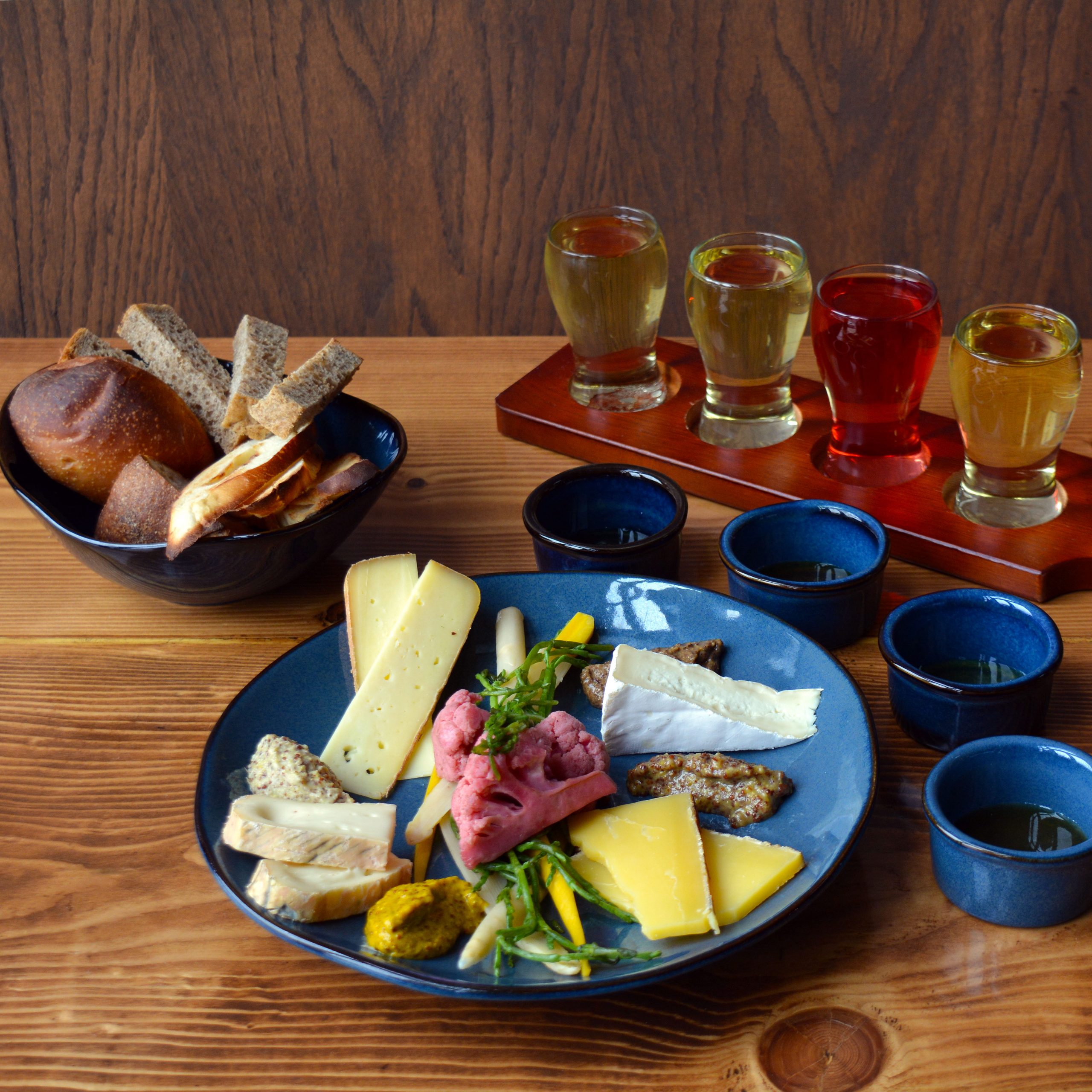 A charcuterie board with varieties of bread, cheese, honey, pickled vegetables, and artisanal mustards, with a flight of cider on the side