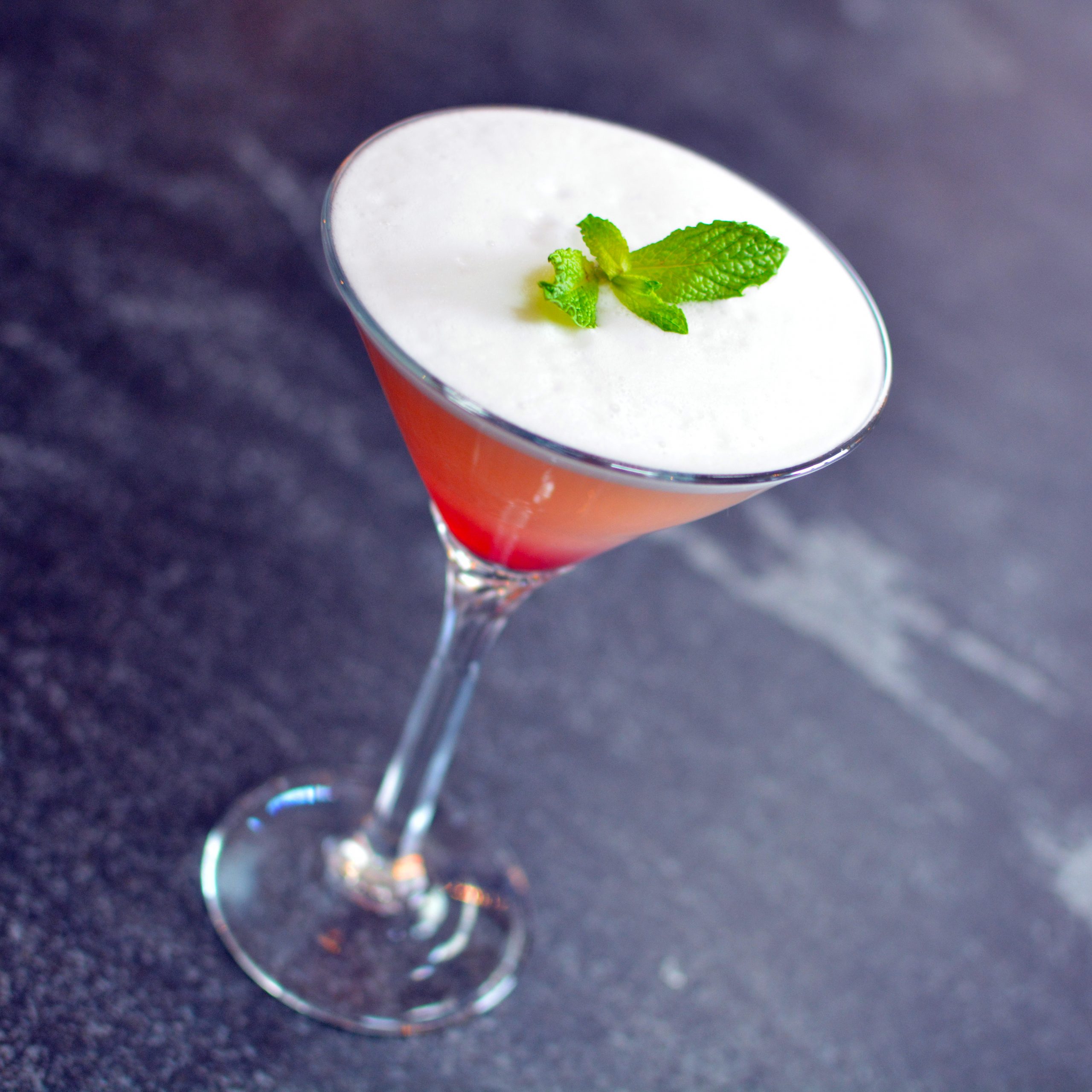 A photo of a red craft cocktail in a martini glass, topped with egg whites and a sprig of mint